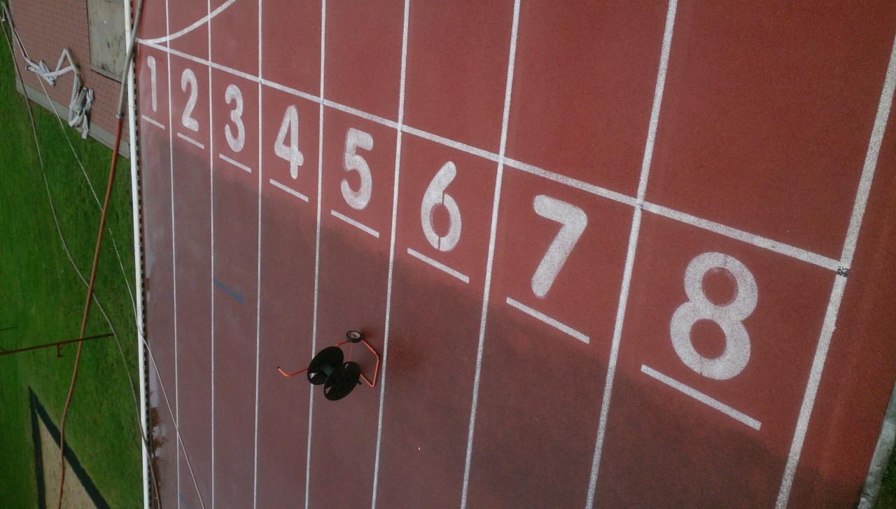 Widok z góry na czerwoną bieżnię lekkoatletyczną z białymi numerami torów od 1 do 8; na torze numer 6 leżą czarne słuchawki nauszne.