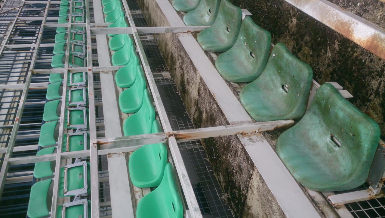 Rzędy brudnych, zielonych plastikowych siedzeń na trybunach sportowych, z widocznymi śladami wilgoci i rdzy na metalowej konstrukcji.