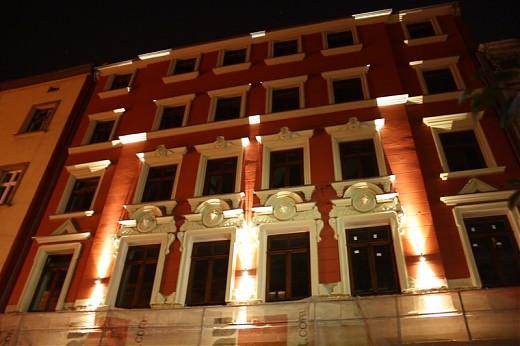 Oświetlona elewacja kamienicy w nocy, widoczne lampy elewacyjne podkreślające architekturę okien i gzymsów.