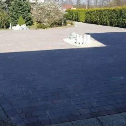 Częściowo zacieniony podjazd z ciemnej kostki brukowej, z widocznymi stertami kostki do ułożenia i fragmentem kratki ściekowej w rogu.