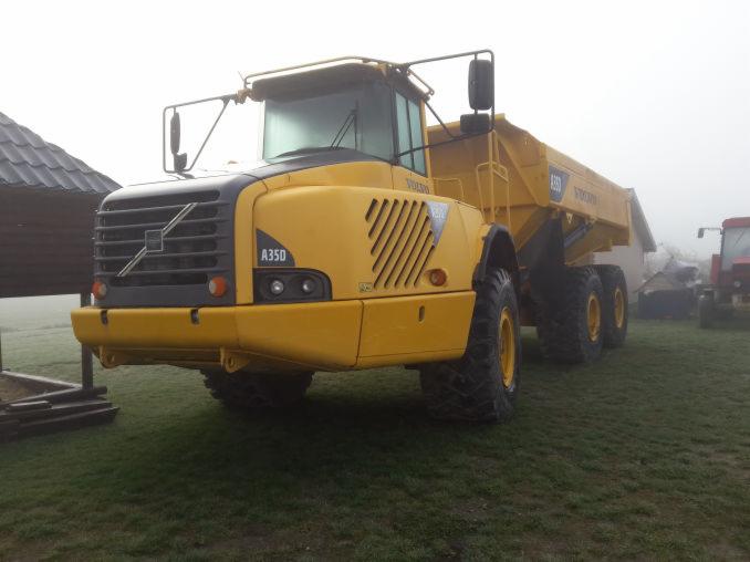 Żółta wywrotka Volvo A35D stoi na trawie w mglisty dzień, z widocznym fragmentem dachu budynku i czerwonym traktorem w tle.