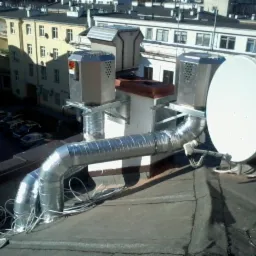 Widok z góry na dachu budynku z zainstalowanymi elementami wentylacji i anteną satelitarną, z widocznymi przewodami i elementami mocującymi.