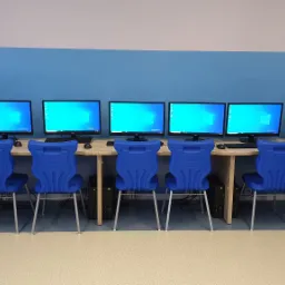 Sześć identycznych stanowisk komputerowych z monitorami AOC, klawiaturami, myszami i niebieskimi krzesłami w jasnym pomieszczeniu biurowym lub pracowni komputerowej.