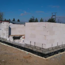 Wznoszenie ścian fundamentowych z bloczków Solbet na budowie domu murowanego, widoczne zabezpieczenie przeciwwilgociowe, w tle drzewa i błękitne niebo.