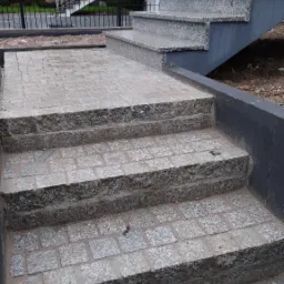 Schody zewnętrzne z kostki granitowej prowadzące do budynku, widok z dołu, z elementami architektury w tle.