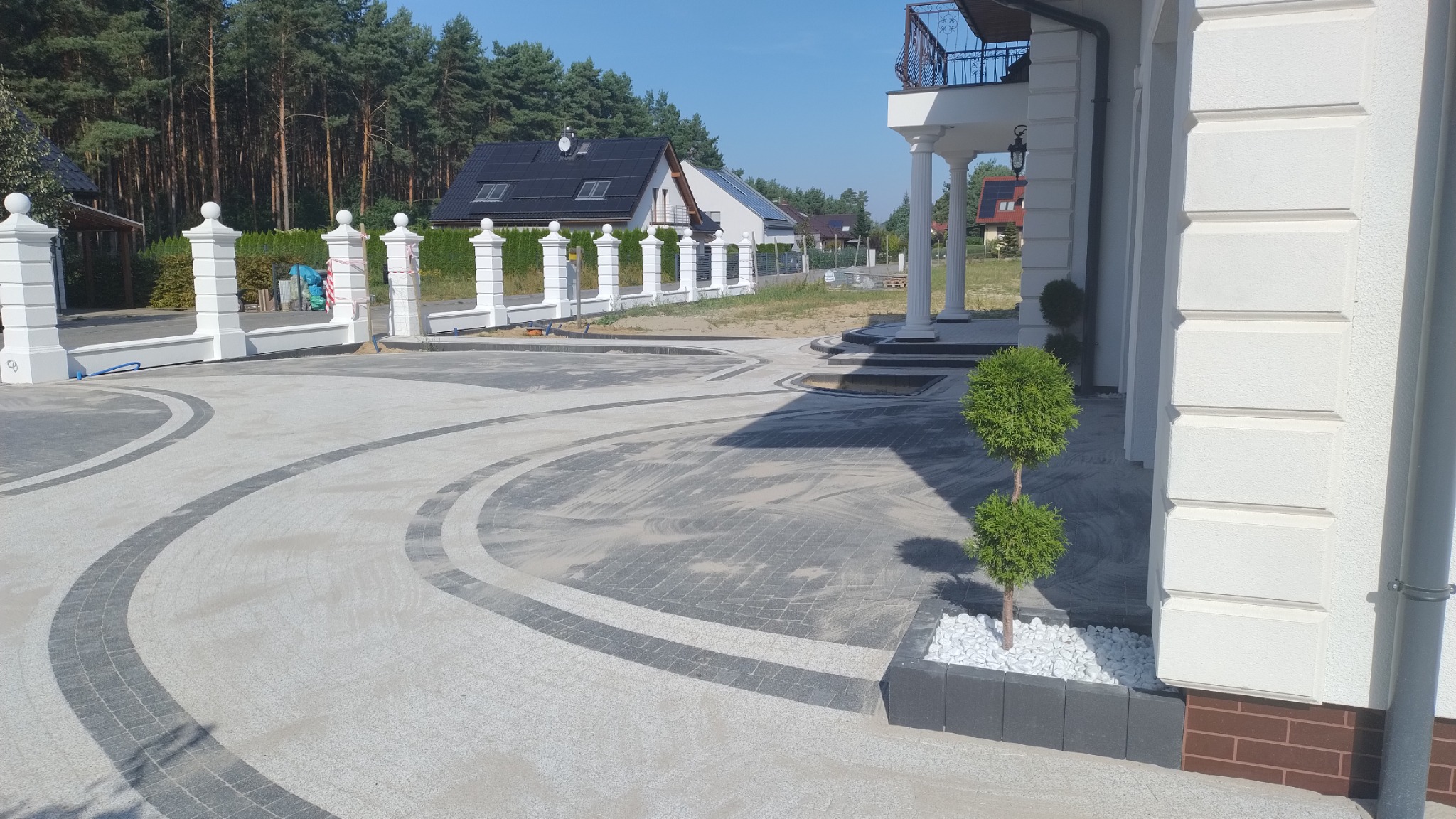 Nowo ułożony podjazd z kostki brukowej w odcieniach szarości, z dekoracyjnymi obramowaniami z ciemniejszej kostki, prowadzący do białego domu z kolumnami i ozdobnym balkonem. Widoczne młode drzewko...