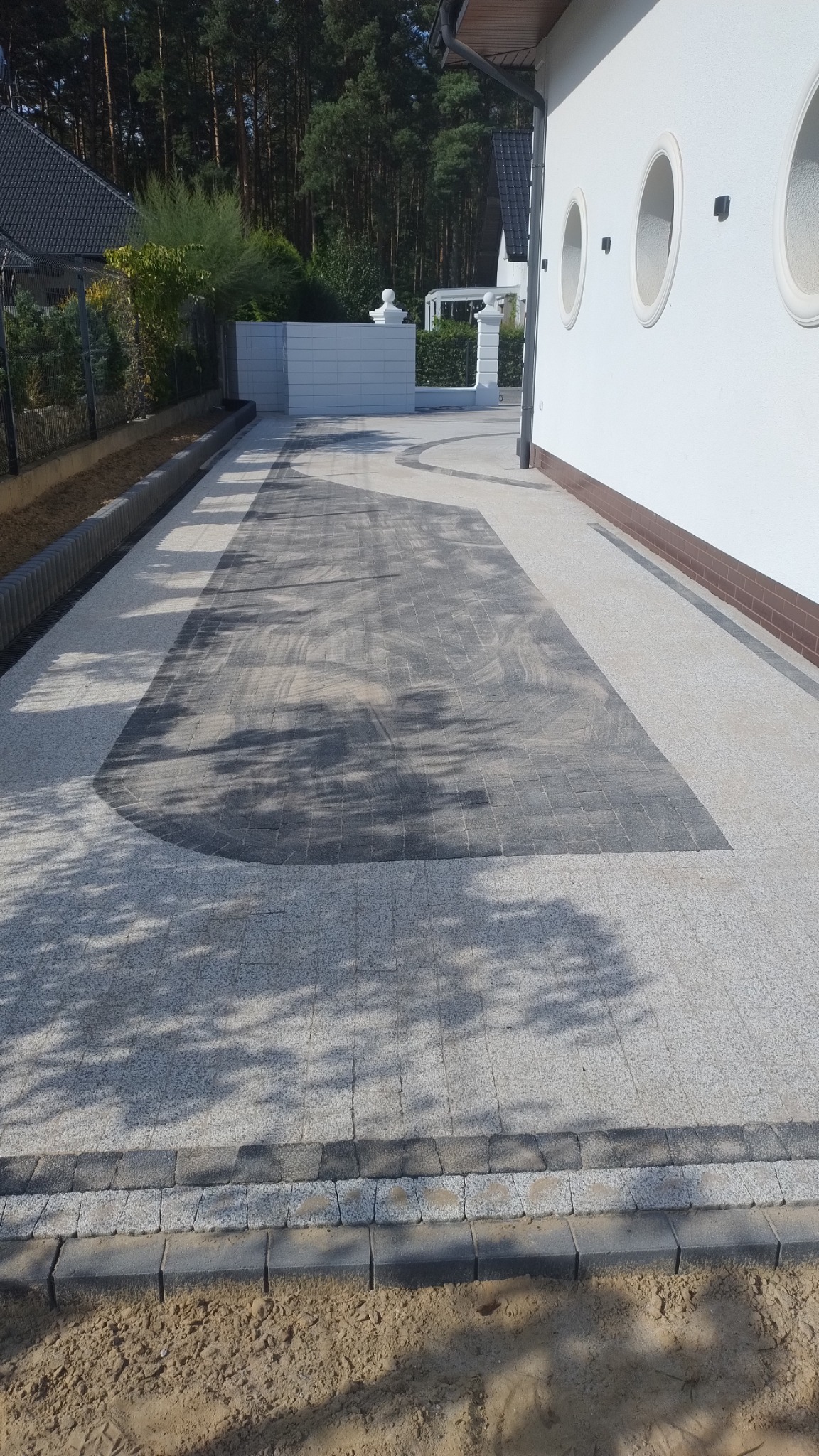 Ułożony podjazd z kostki w dwóch odcieniach szarości, z ciemniejszym prostokątnym wzorem na środku. Krawężnik z kostki oddziela podjazd od piasku. Biały budynek z okrągłymi oknami w tle.