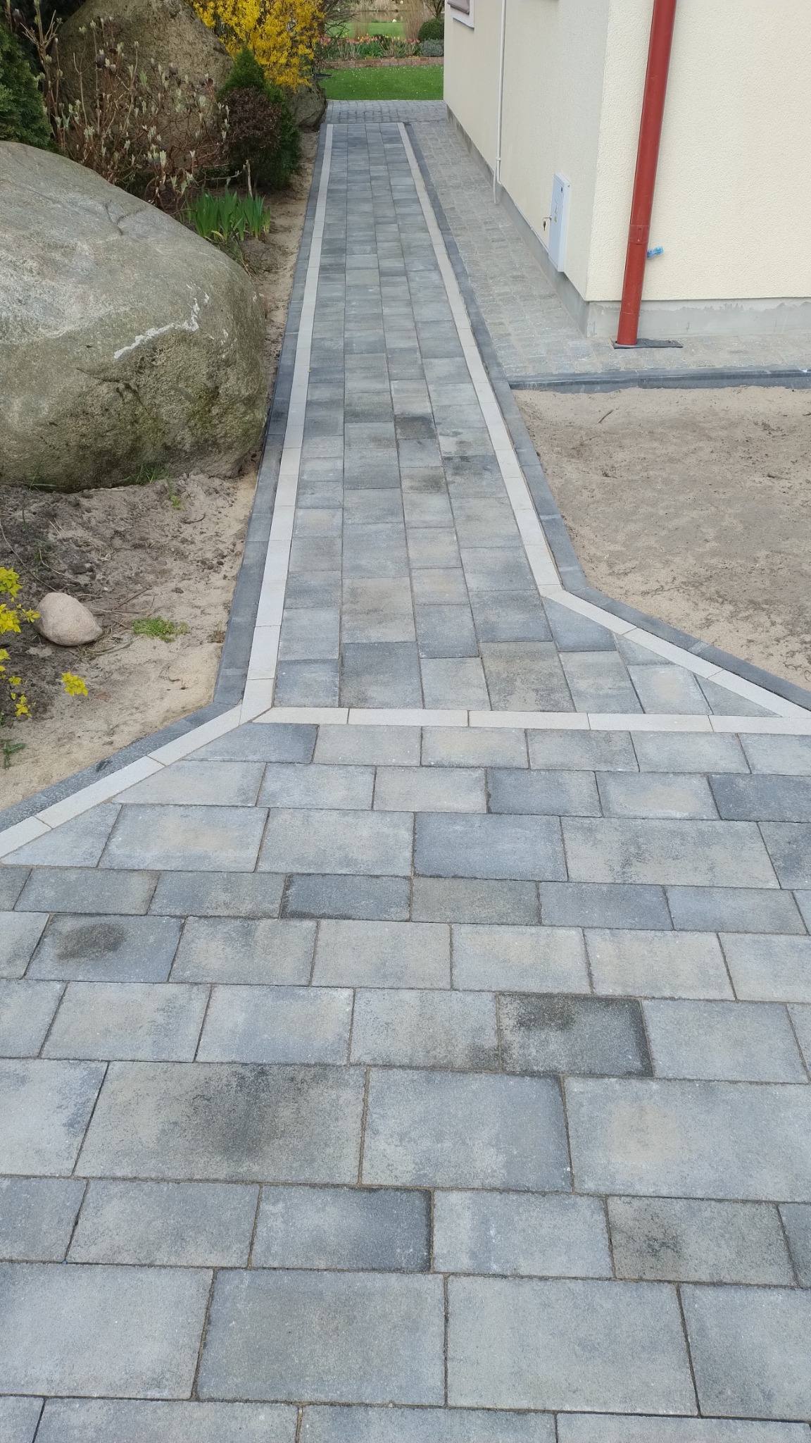 Ścieżka z szarej kostki brukowej z jasnymi obrzeżami prowadząca wzdłuż budynku, obok duży kamień i kwitnące krzewy.