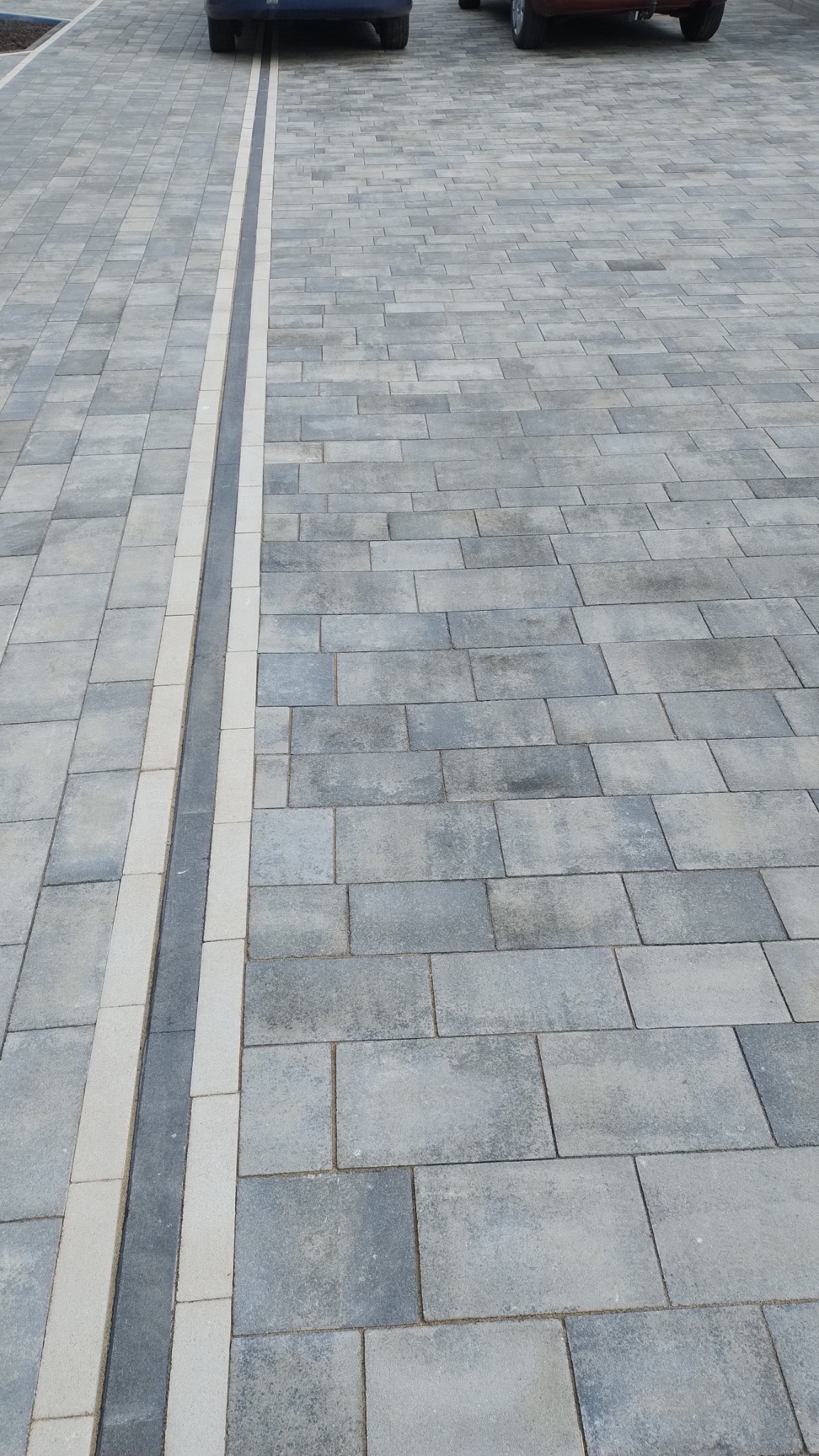 Szara kostka brukowa ułożona na podjeździe z liniowym odpływem wody obramowanym jasnymi kostkami, widoczne fragmenty samochodów w tle.