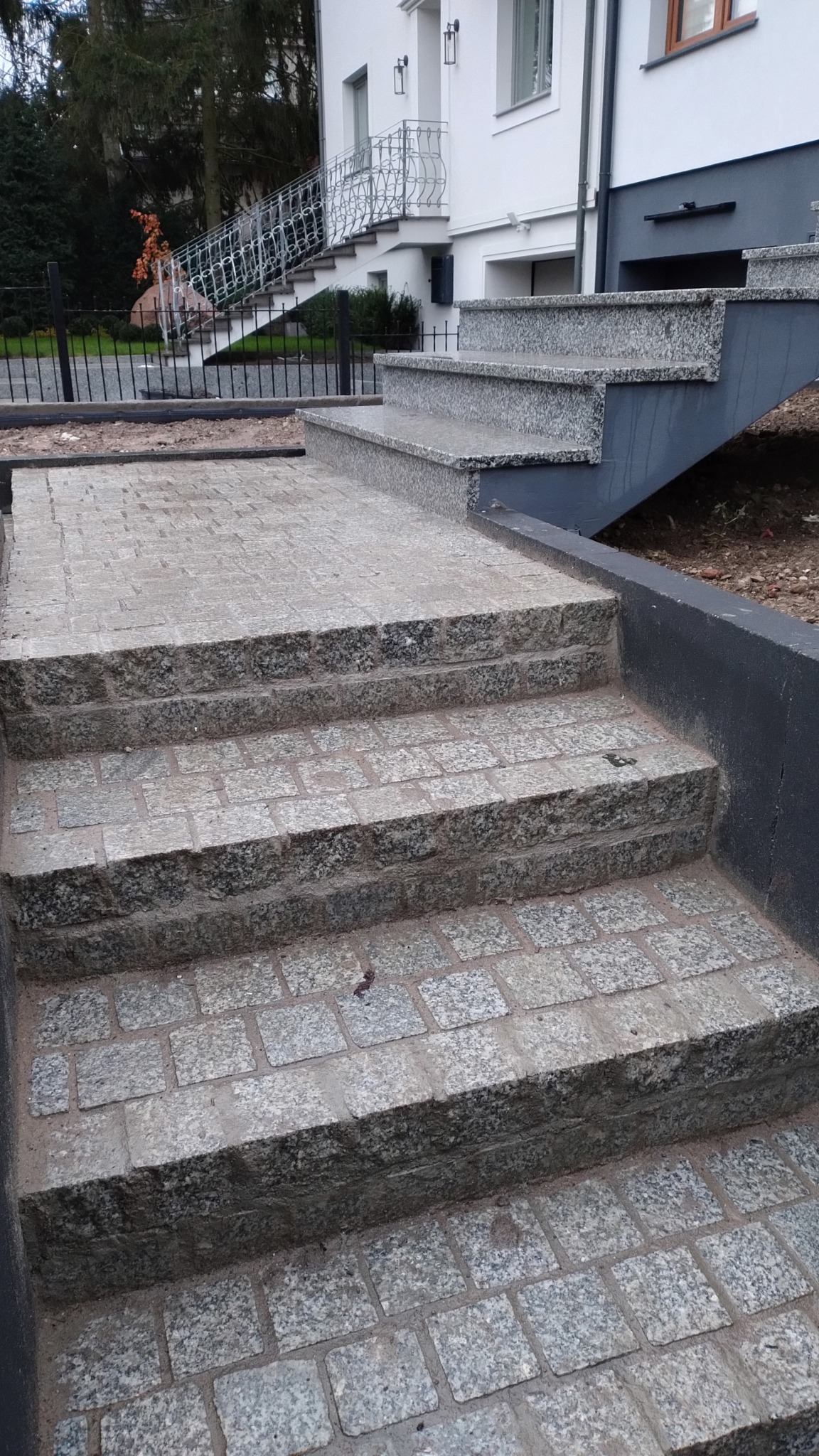 Schody zewnętrzne z kostki granitowej prowadzące do budynku, widok z dołu, z elementami architektury w tle.