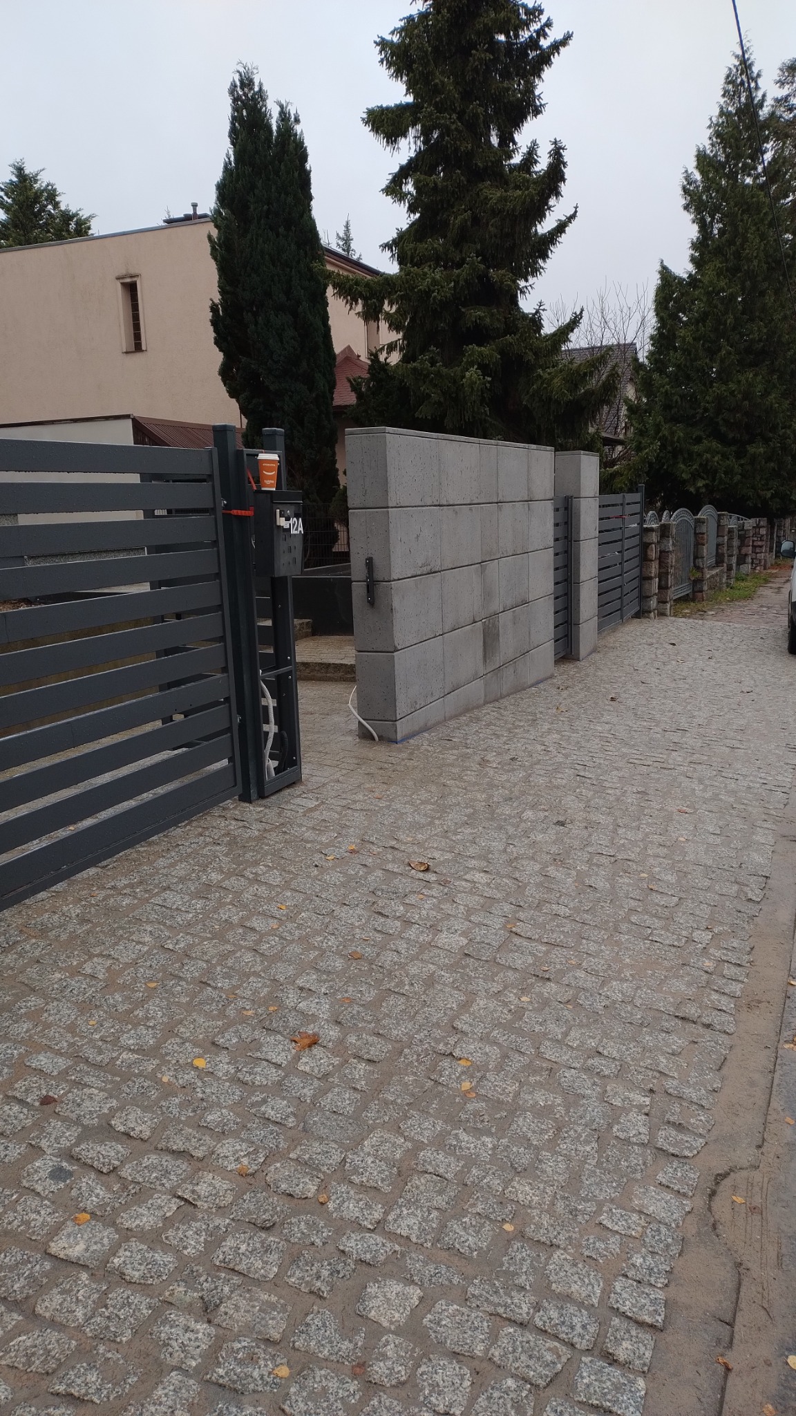 Ułożona kostka granitowa prowadząca do nowoczesnej posesji z szarą bramą i betonowym ogrodzeniem modułowym; na bramie kubek po kawie.