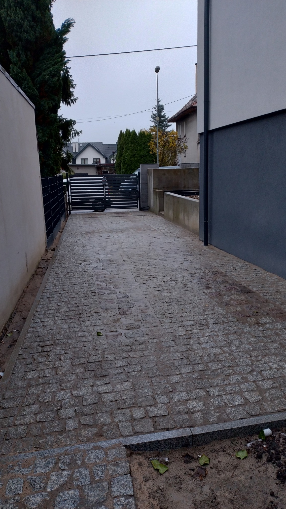 Ułożona nawierzchnia z kostki granitowej prowadząca do bramy wjazdowej, widok z perspektywy wjazdu, z elementami krajobrazu i zabudowy w tle.
