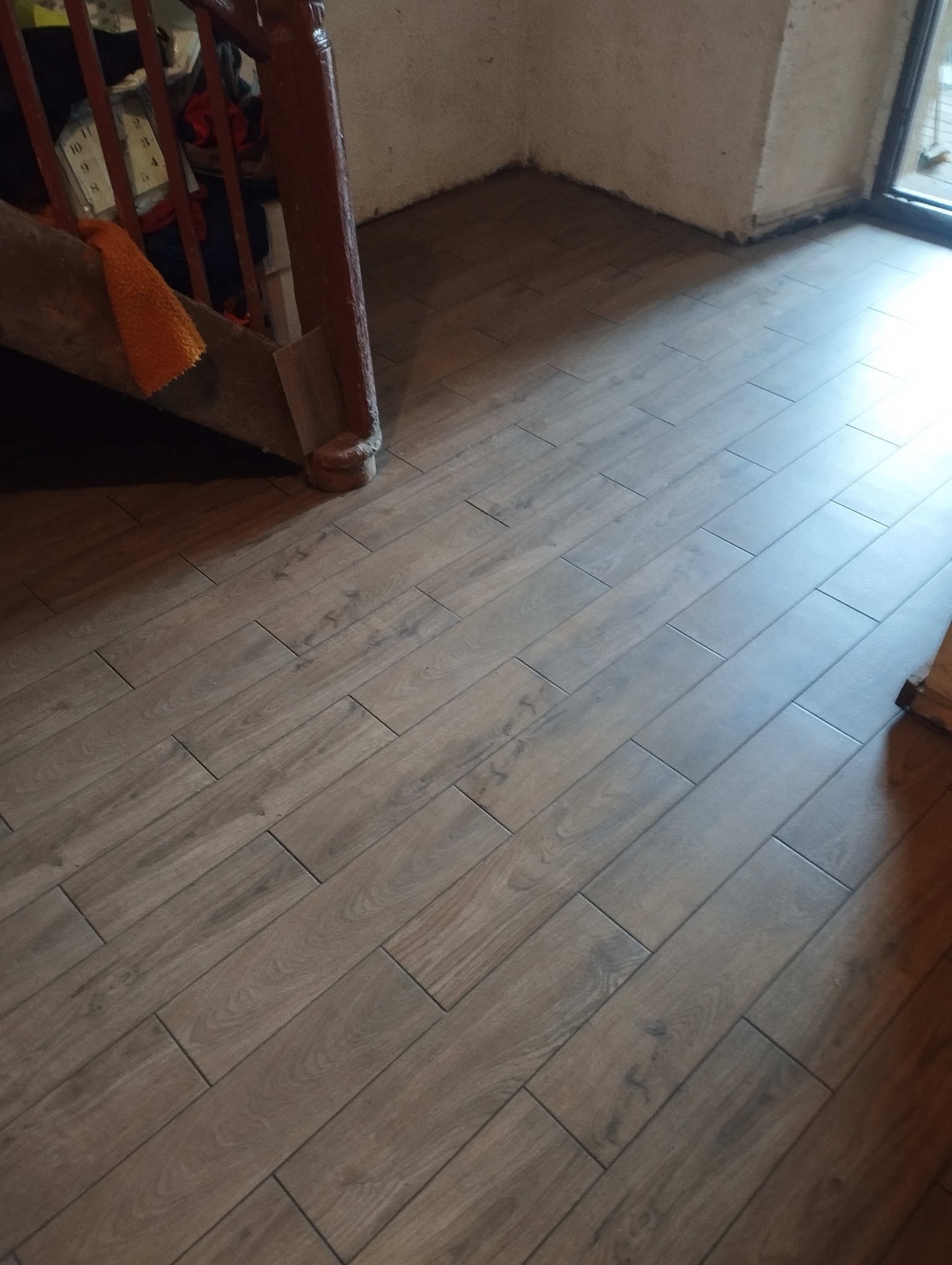 Ukończona podłoga z płytek imitujących drewno, ułożonych w jodełkę, widoczna przy schodach i ścianie. Jasne, naturalne światło podkreśla wzór.