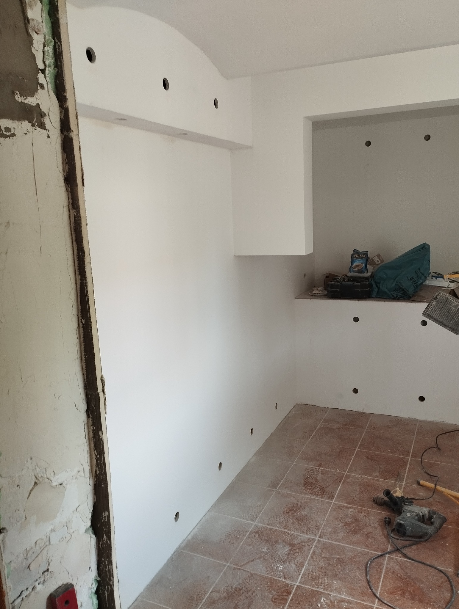 Wnętrze po szpachlowaniu i malowaniu ścian na biało, widoczne otwory pod oświetlenie, fragment starej futryny i narzędzia na podłodze z płytek.