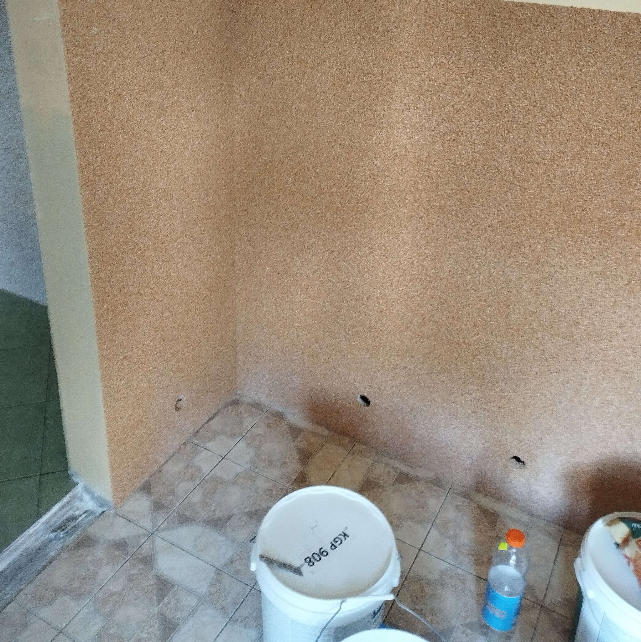 Ściana pokryta tapetą natryskową w odcieniach beżu, z widocznymi puszkami po farbie na podłodze wyłożonej płytkami. Widoczne otwory w ścianie.