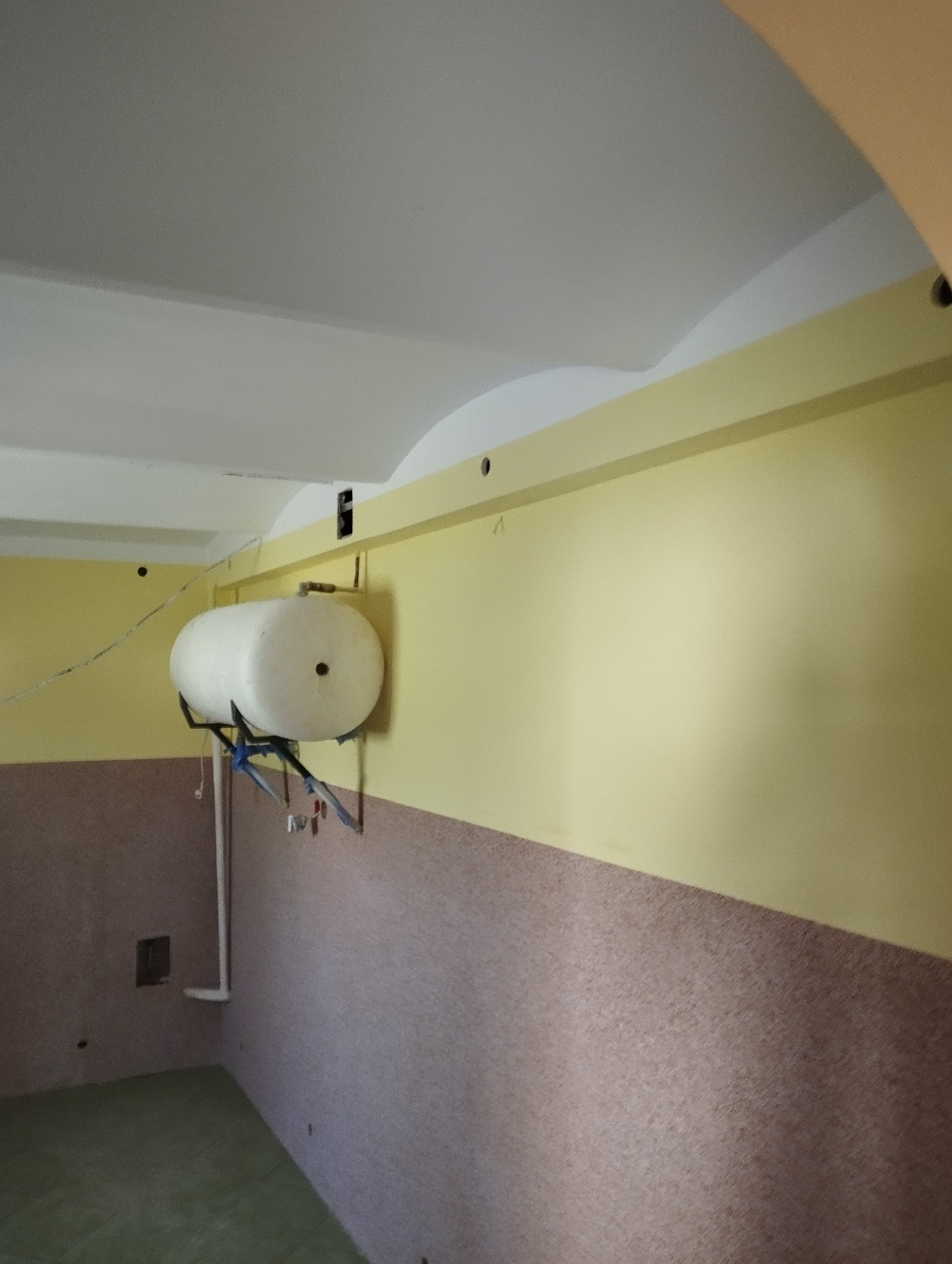 Wnętrze pokoju z żółtymi ścianami, wykończonym sufitem i zamontowanym zbiornikiem na ścianie. Widoczne elementy instalacji i wykończenia ścian.