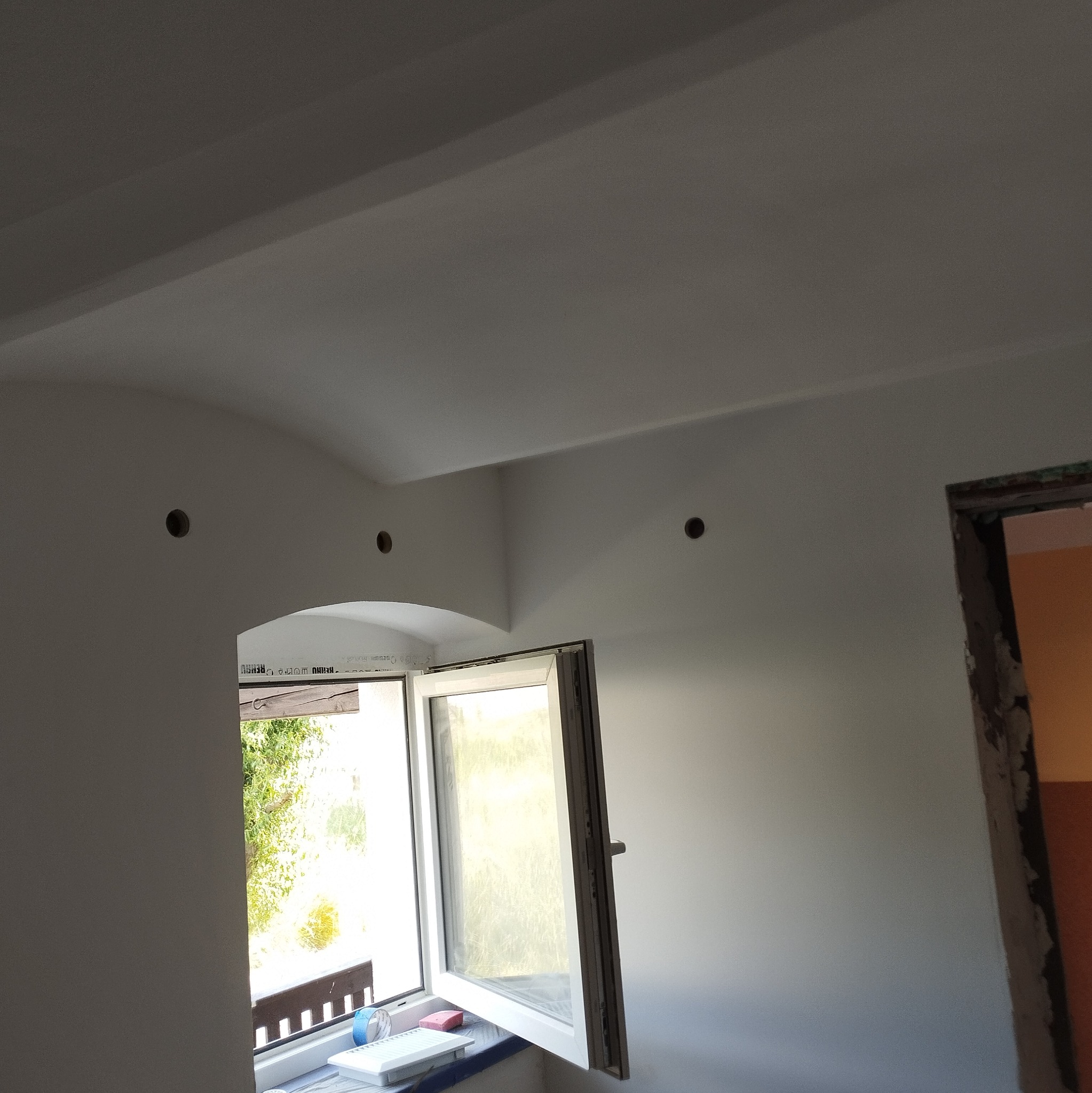 Wnętrze po remoncie: gładkie, białe ściany z otworami na oświetlenie, otwarte okno z widokiem na zieleń. Wykończone, jasne pomieszczenie z elementami przygotowanymi do dalszych prac.