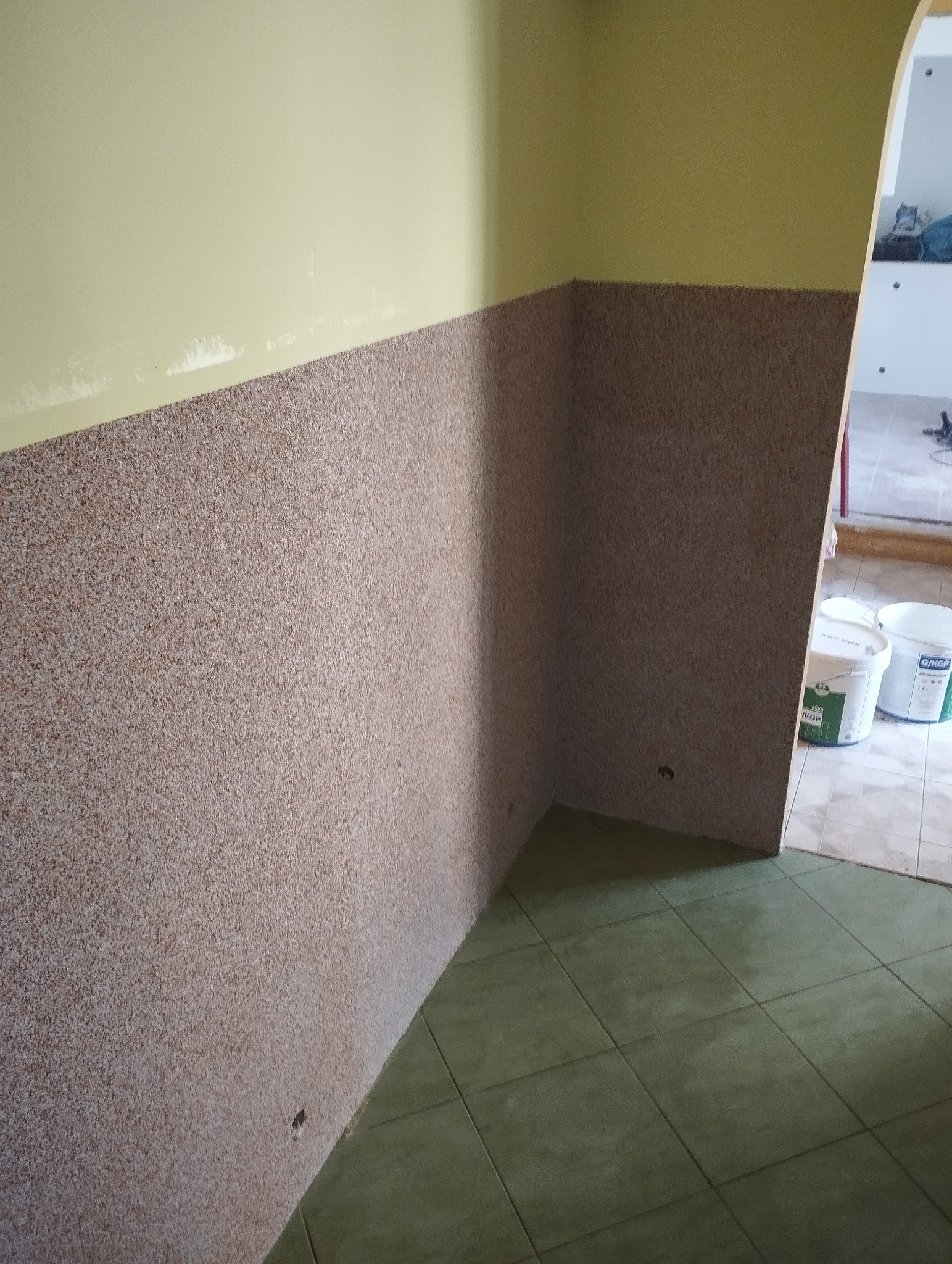 Wykończona ściana z dekoracyjnym tynkiem w odcieniach brązu i beżu, kontrastująca z górną częścią ściany pomalowaną na jasnożółty kolor. Widoczne wiaderka z farbą w tle.