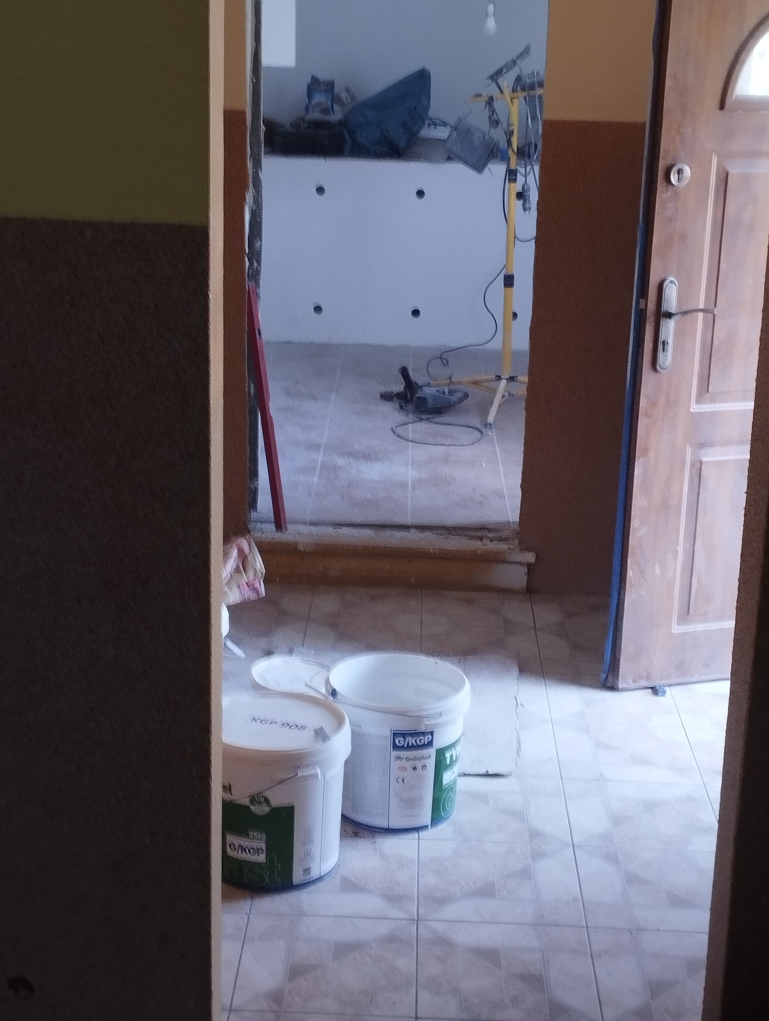 Wnętrze w trakcie remontu: widoczne wiadra z zaprawą, narzędzia i oświetlenie. Ściany przygotowane do dalszych prac wykończeniowych, widoczne otwory pod instalacje.