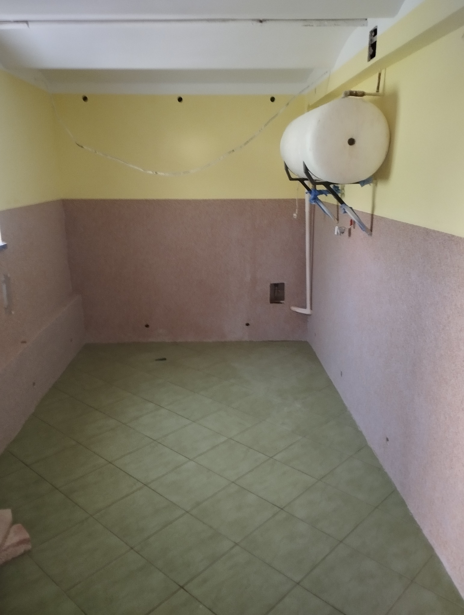 Wnętrze pomieszczenia z wykończonymi ścianami do połowy wysokości i zieloną podłogą z płytek. Na ścianie zamontowany biały zbiornik na wodę.