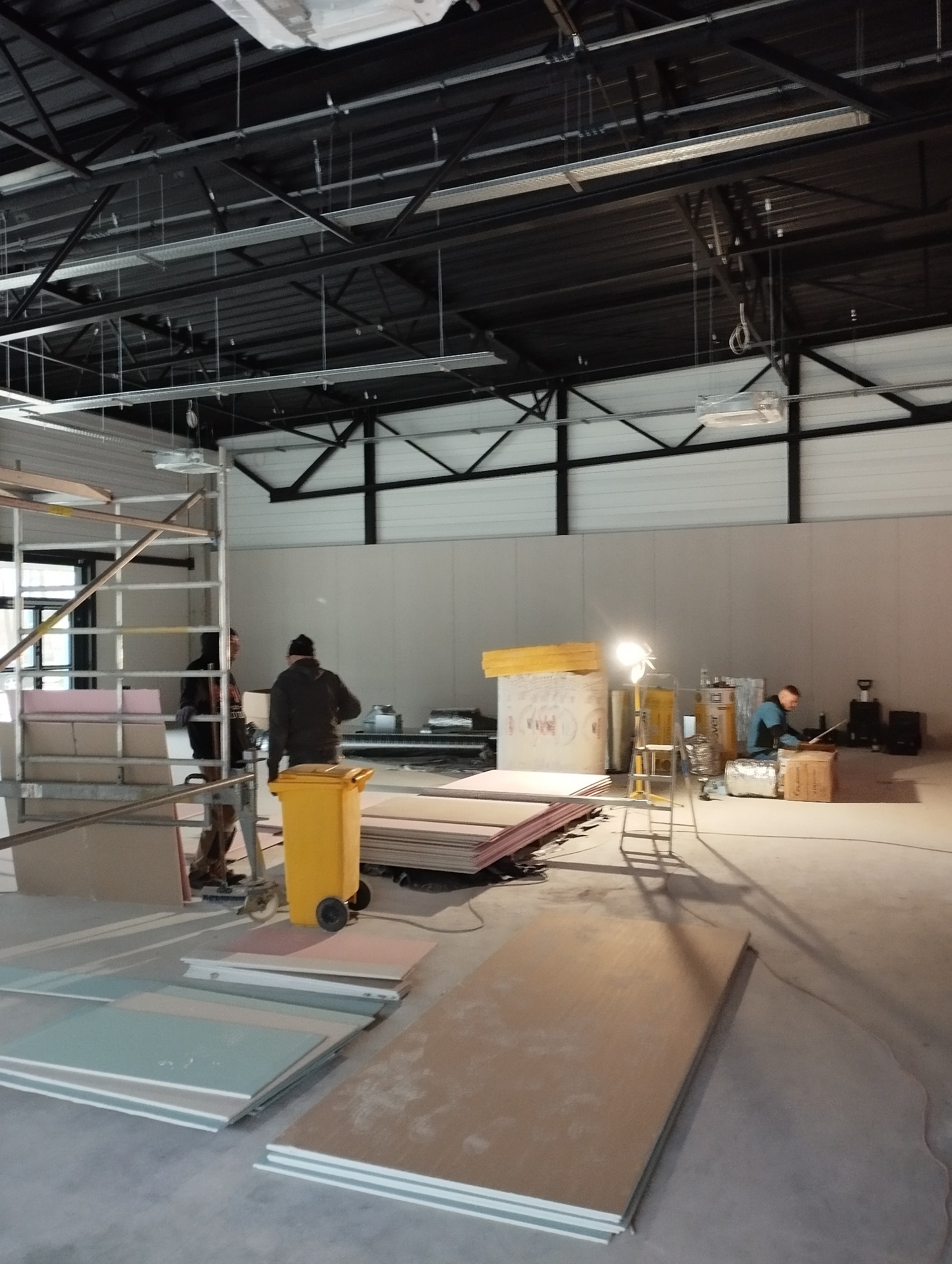 Montaż płyt w hali: pracownicy przy rusztowaniu, stosy płyt, oświetlenie budowlane. Widoczne elementy konstrukcji dachu i ścian. Żółty pojemnik na odpady budowlane.