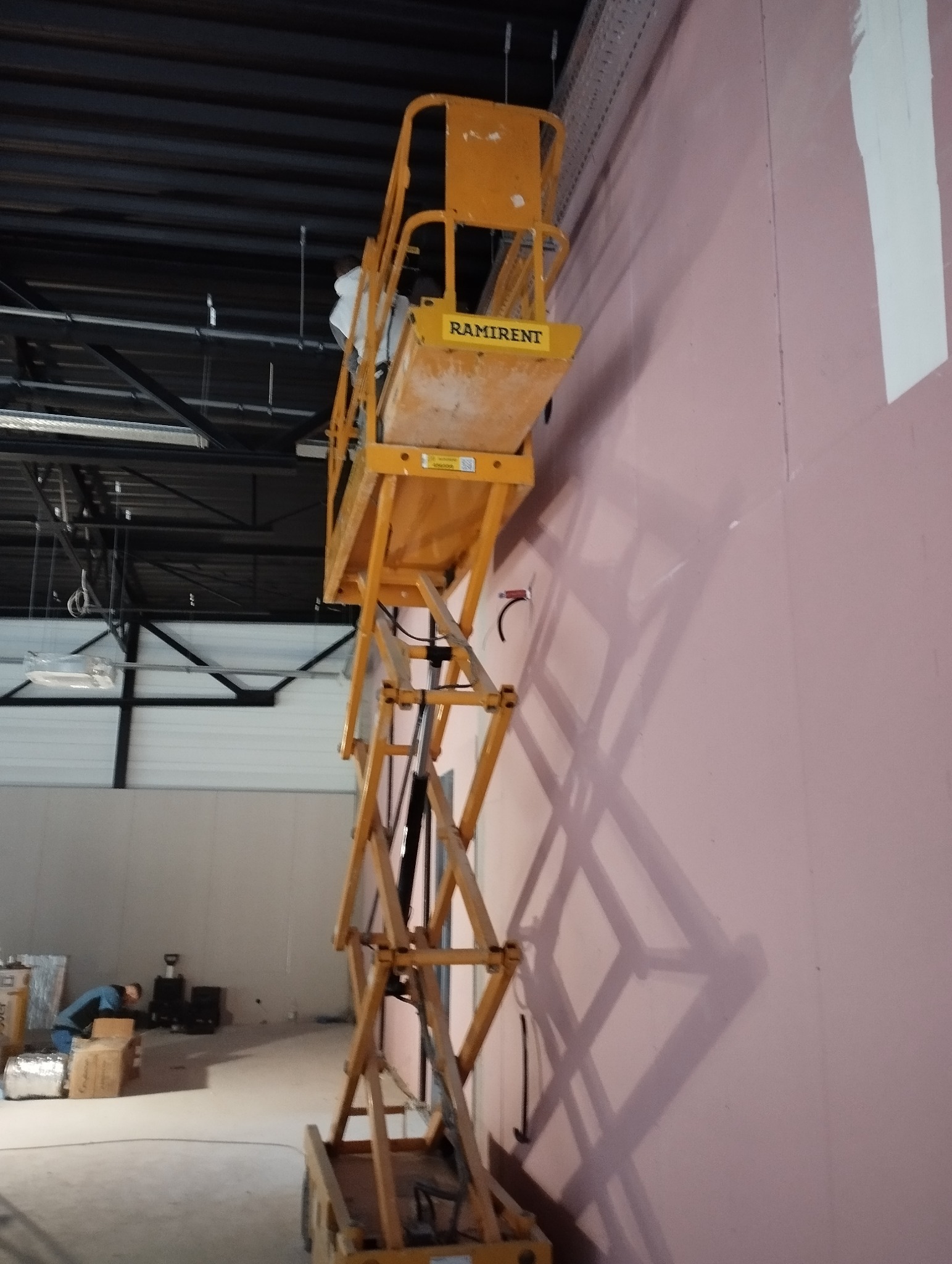 Montaż płyt na ścianie z podnośnika. Widoczna surowa ściana z płyt, podnośnik nożycowy RAMIRENT i pracownik na wysokości. Hala z widoczną konstrukcją sufitu.
