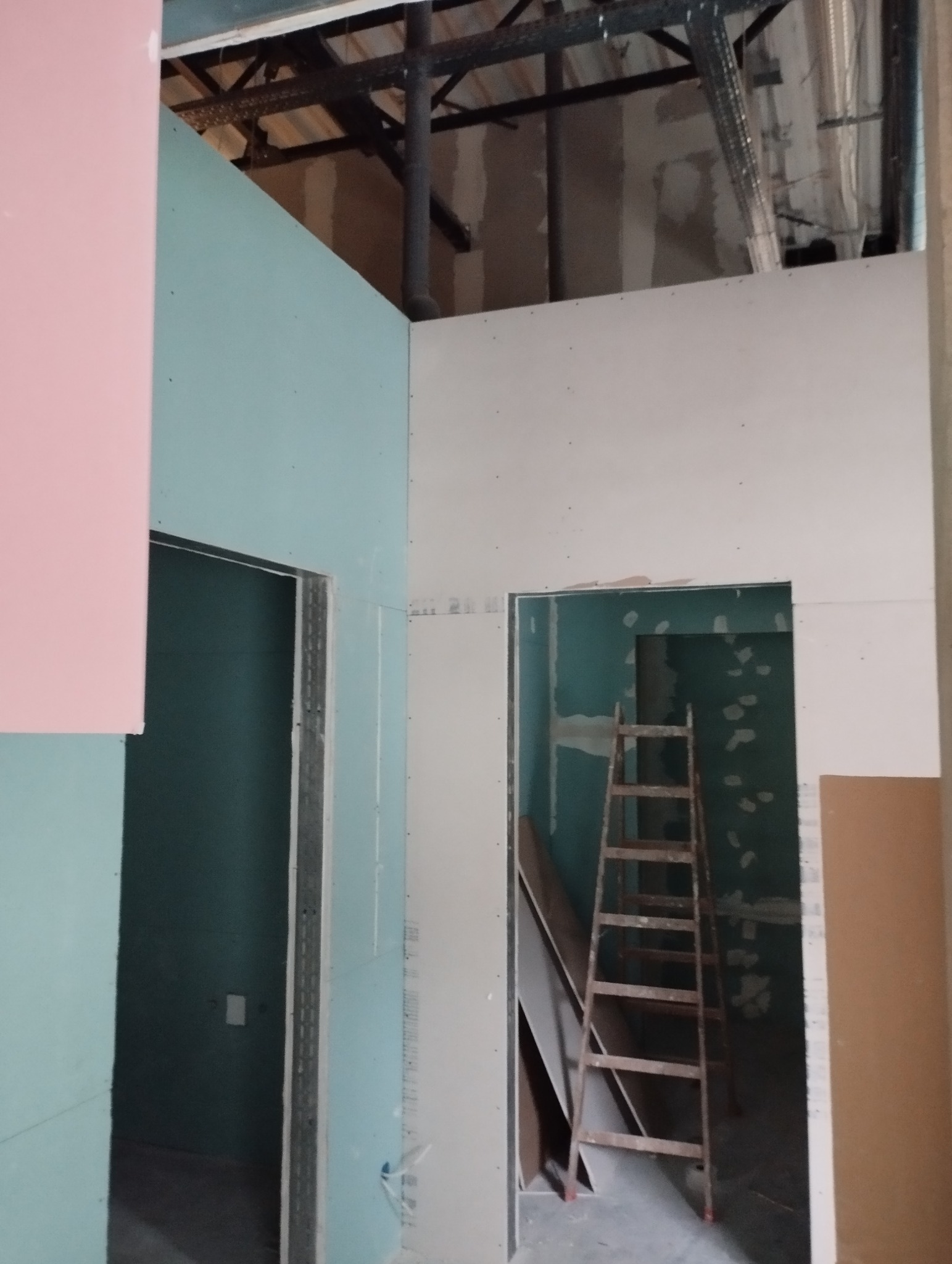 Montaż ścian działowych z płyt gipsowo-kartonowych w surowym wnętrzu. Widoczna drabina i konstrukcja sufitu. Ściany w kolorach: różowy, zielony i biały.