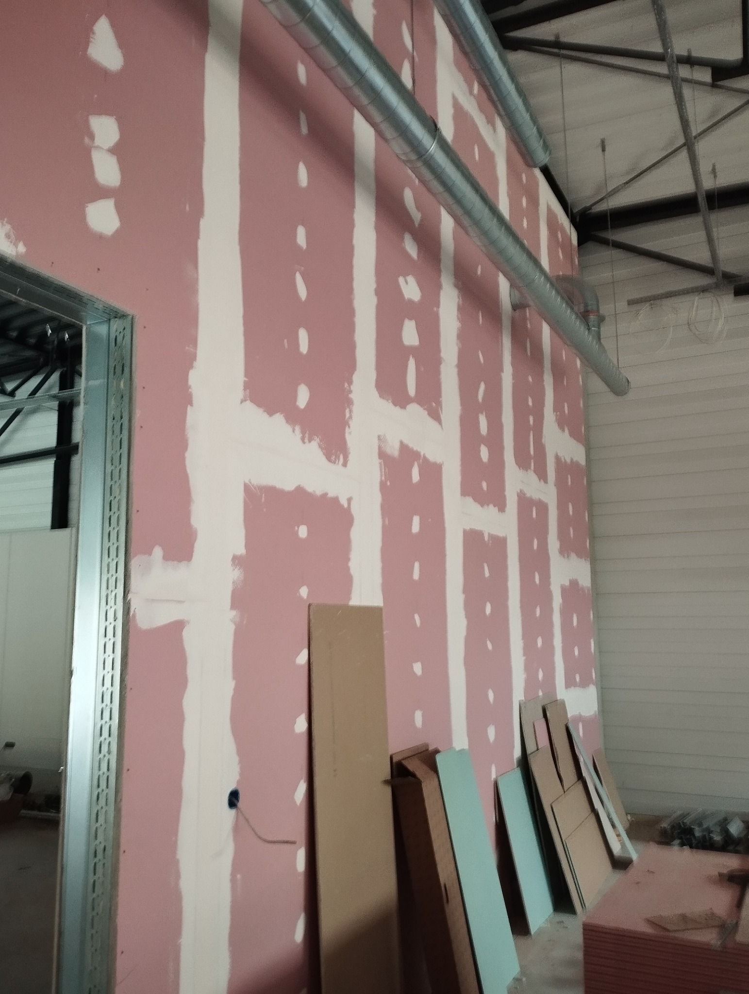 Ściana z płyt karton-gips, częściowo zaszpachlowana, widoczne metalowe rury wentylacyjne pod sufitem i stos płyt przy ścianie. W tle widoczna konstrukcja dachu.