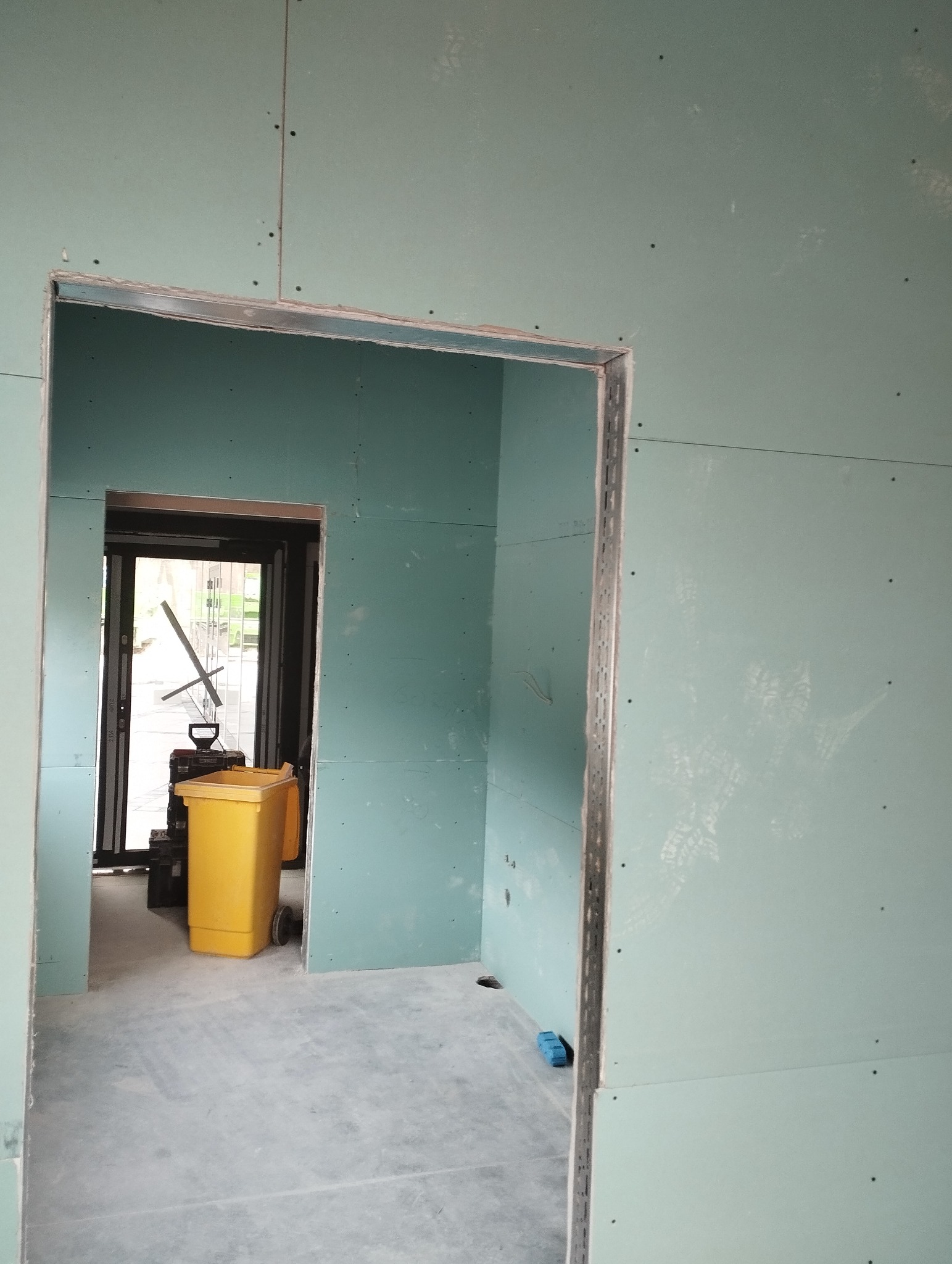 Ściana z płyt karton-gips z wyciętym otworem na drzwi, widoczny żółty pojemnik na śmieci i narzędzia w tle. Prace wykończeniowe w surowym wnętrzu.