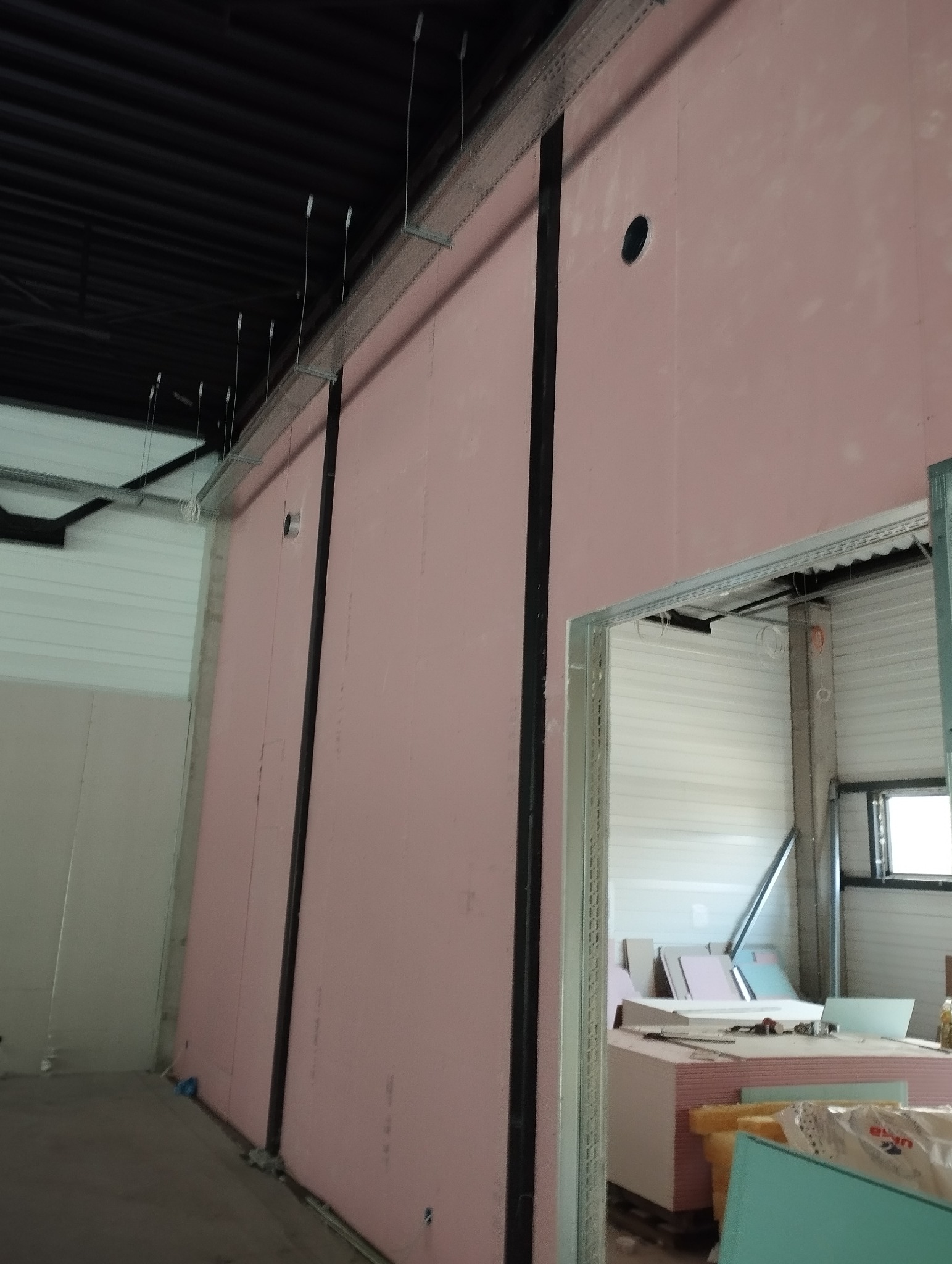 Montaż różowych płyt gipsowo-kartonowych na metalowym stelażu w hali. Widoczne otwory na instalacje i elementy konstrukcyjne sufitu. Prace wykończeniowe w toku.