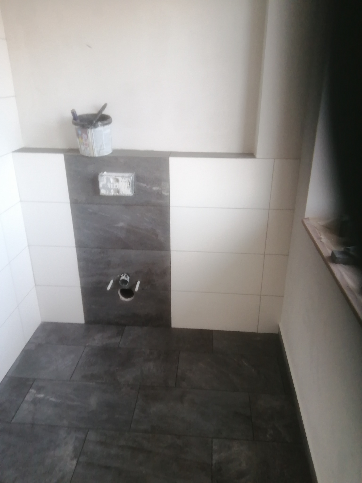 Wykończona łazienka z szarymi płytkami podłogowymi i białymi/szarymi płytkami ściennymi. Widoczna instalacja podtynkowa WC i wiadro z narzędziami.