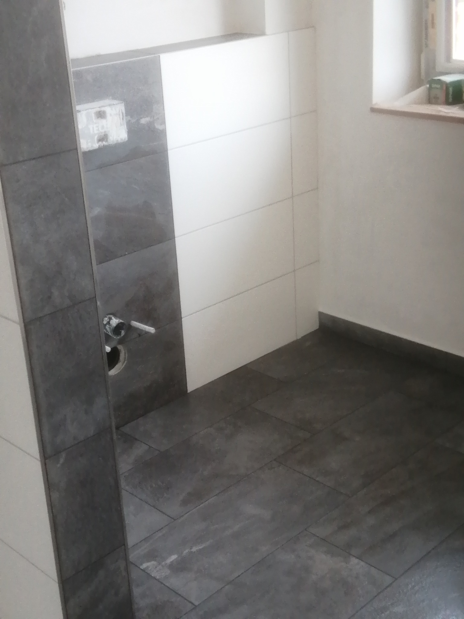 Wykończona łazienka z szarymi płytkami podłogowymi i częściowo wykończonymi ścianami w odcieniach bieli i szarości. Widoczny fragment instalacji wodnej.