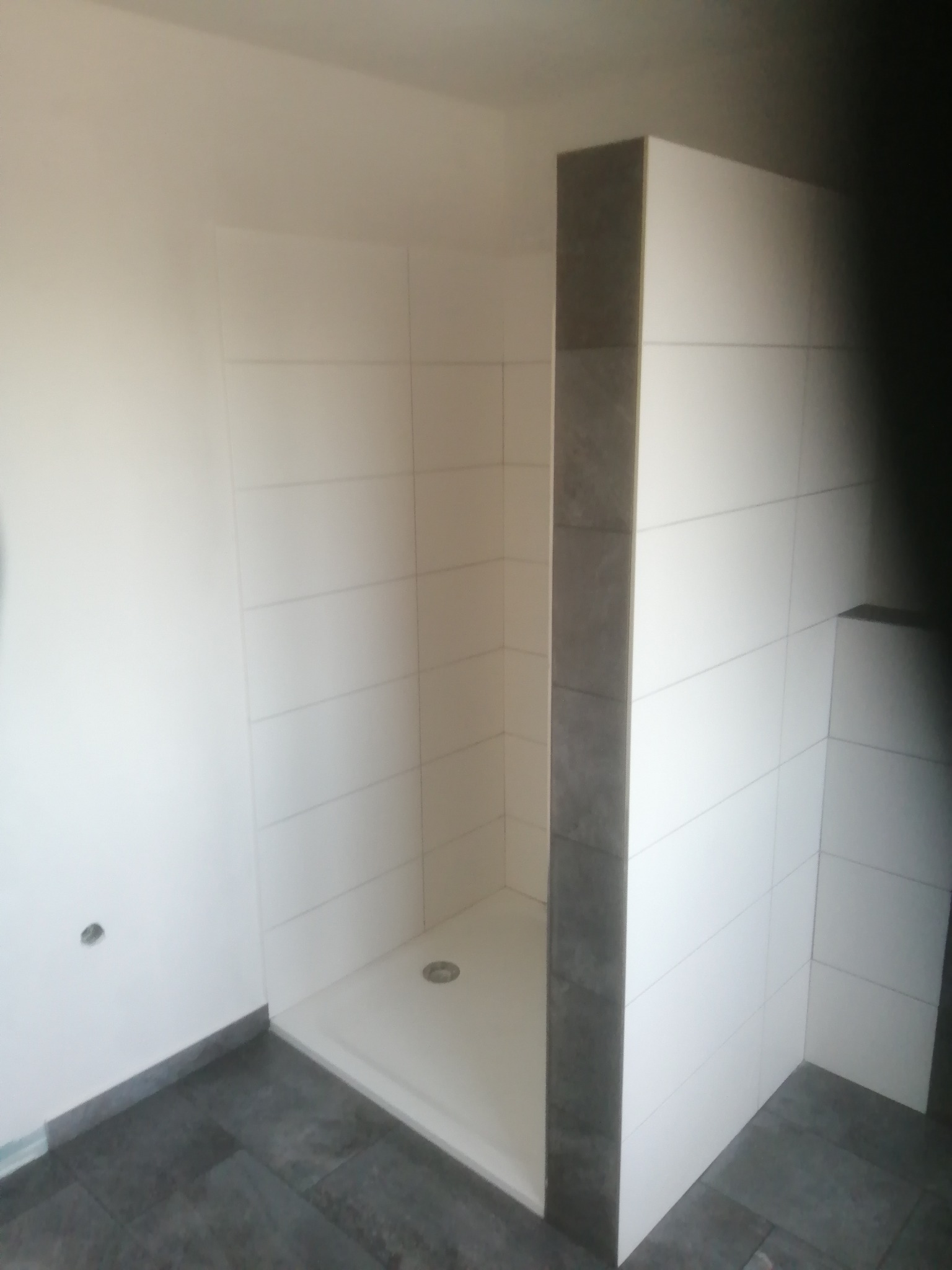 Nowoczesna kabina prysznicowa z białymi i szarymi płytkami, widoczny brodzik i fragment podłogi z szarych płytek. Minimalistyczny design, surowe wykończenie.