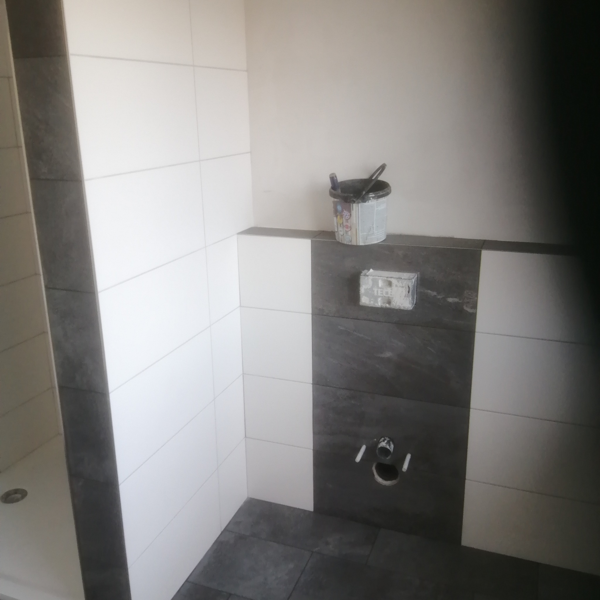 Nowoczesna łazienka w trakcie wykańczania: biało-szare płytki, widoczne przyłącza i wiadro z narzędziami. Minimalistyczny design z akcentem na detale.