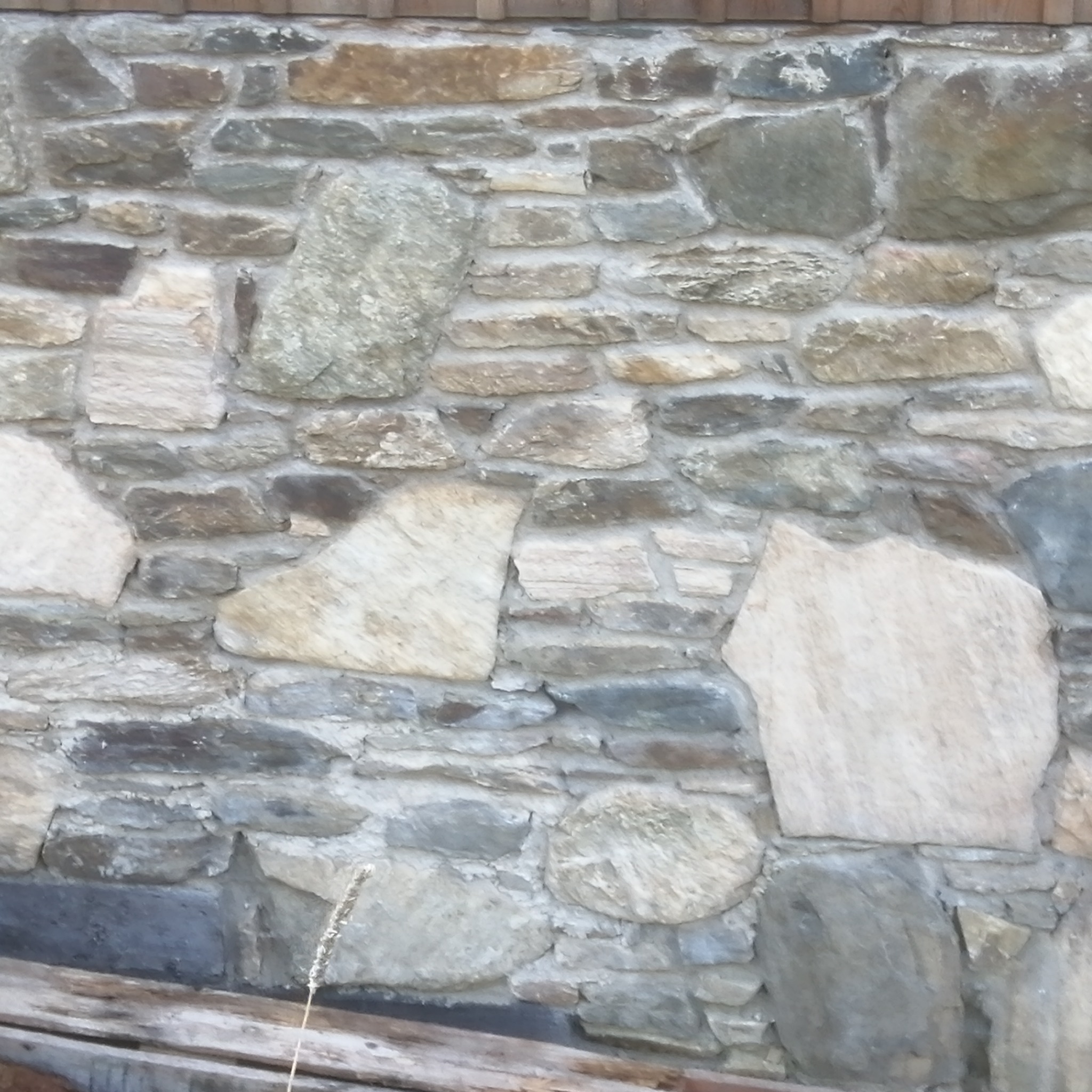 Kamienna ściana z nieregularnych, szarych i beżowych kamieni, tworząca rustykalną teksturę. Widoczne spoiny między kamieniami i fragment drewnianej konstrukcji u dołu.