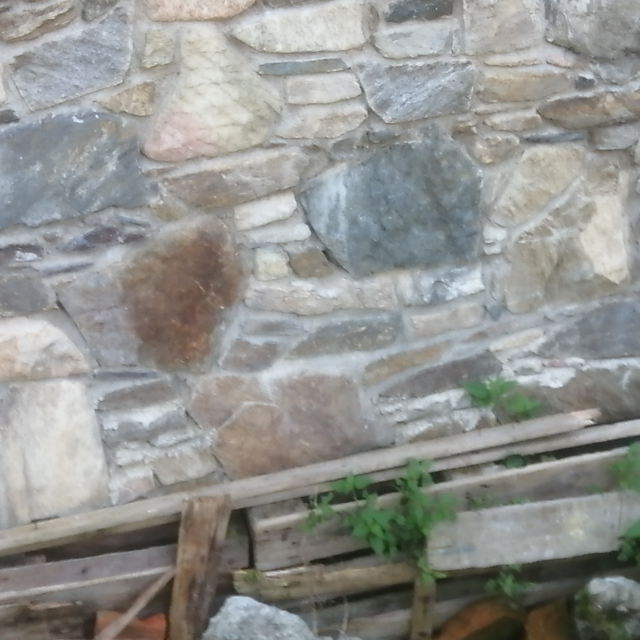 Kamienna ściana z nieregularnych, szarych i beżowych kamieni, częściowo zasłonięta przez drewniane elementy i zieleń. Nierówna faktura kamienia, naturalne oświetlenie.