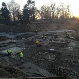 Siatka zbrojeniowa ułożona na mokrym podłożu budowy, pracownicy w żółtych kamizelkach i kaskach, widoczne w oddali drzewa i zachodzące słońce.
