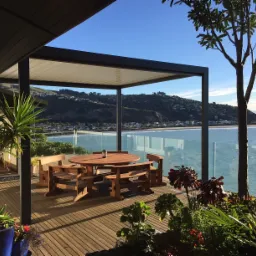 Pergola z drewnianym stołem i krzesłami na tarasie z widokiem na morze, otoczona roślinnością, w słoneczny dzień.