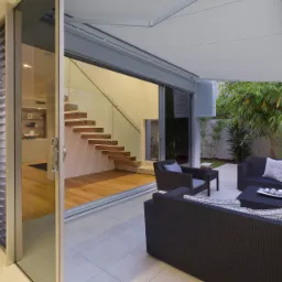 Nowoczesny taras z meblami rattanowymi pod rozkładaną markizą, widok na wnętrze domu przez przesuwne szklane drzwi z aluminiową ramą.