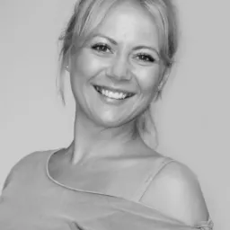 Portret uśmiechniętej blondynki z włosami upiętymi w kok, ubrana w luźną bluzkę z odsłoniętym ramieniem, na jasnym tle.