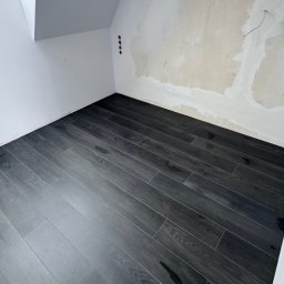 Firma ogolno budowlana Miroslaw Gros - Ukończona podłoga z ciemnych, drewnopodobnych paneli w pokoju na poddaszu. Ściany częściowo przygotowane do malowania. Widoczne gniazdka elektryczne.