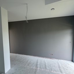 Firma ogolno budowlana Miroslaw Gros - Pomieszczenie po malowaniu ścian na szaro, z widocznymi puszkami elektrycznymi. Podłoga zabezpieczona folią malarską. W tle biały sufit z wystającym przewodem.