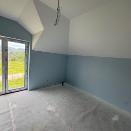 Firma ogolno budowlana Miroslaw Gros - Poddasze po malowaniu ścian na niebiesko, widoczne okno z widokiem na zielone wzgórza, przygotowana podłoga, wystające kable z sufitu. Świeżo po remoncie.