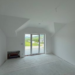 Firma ogolno budowlana Miroslaw Gros - Wnętrze pokoju z dużym oknem balkonowym i instalacją grzewczą w ścianie. Podłoga zabezpieczona folią, ściany i sufit pomalowane na biało. Widok na zewnątrz.