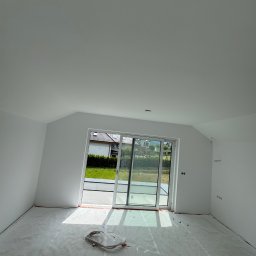 Firma ogolno budowlana Miroslaw Gros - Jasne wnętrze po gładziowaniu z widocznymi kablami na podłodze i dużym oknem tarasowym, wpadające światło dzienne. Ściany i sufit pomalowane na biało.