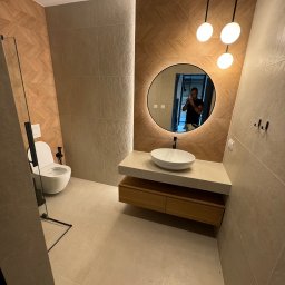 Firma ogolno budowlana Miroslaw Gros - Nowoczesna łazienka z okrągłym lustrem z podświetleniem LED, drewnianą szafką pod umywalką i geometryczną mozaiką na ścianie. Widoczna kabina prysznicowa.