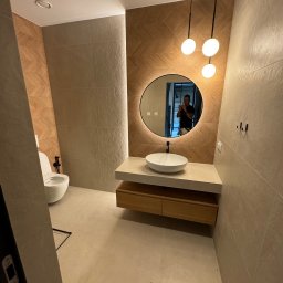 Firma ogolno budowlana Miroslaw Gros - Nowoczesna łazienka z beżowymi płytkami, okrągłym lustrem z podświetleniem LED, wiszącą szafką z umywalką i podwieszaną toaletą. Na ścianie wieszaki.