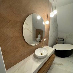 Firma ogolno budowlana Miroslaw Gros - Nowoczesna łazienka z owalnym lustrem, umywalką na blacie z marmuru i wolnostojącą wanną. Na ścianie płytki w jodełkę, a w lustrze odbicie toalety.