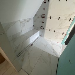 Firma ogolno budowlana Miroslaw Gros - Łazienka w trakcie wykańczania: białe płytki imitujące marmur na ścianach i podłodze, system poziomowania płytek, widoczny odpływ liniowy, skos dachu.