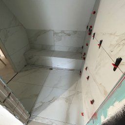 Firma ogolno budowlana Miroslaw Gros - Wykończenie łazienki: białe płytki imitujące marmur na ścianach i podłodze, z systemem poziomowania płytek. Widoczny odpływ liniowy i fragment przygotowanej ściany.